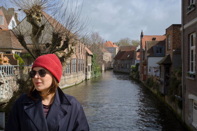 Una mujer con gorro rojo y gafas oscuras mira hacia un lado ante un canal entre casas de Brujas.