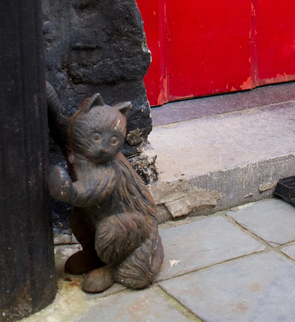 Una pequeña escultura de gato se apoya en un poste oscuro junto al umbral desgastado de una puerta roja.