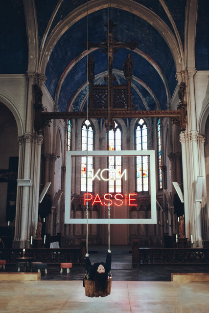 Una persona cuelga boca abajo bajo el neón «KOM PASSIE», entre arcos y vidrieras de una iglesia.