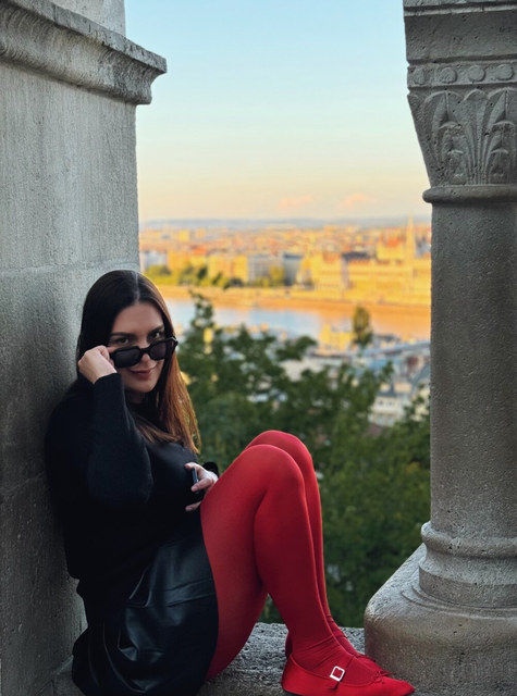 Una mujer con gafas oscuras y medias rojas se sienta en un mirador de piedra sobre Budapest.