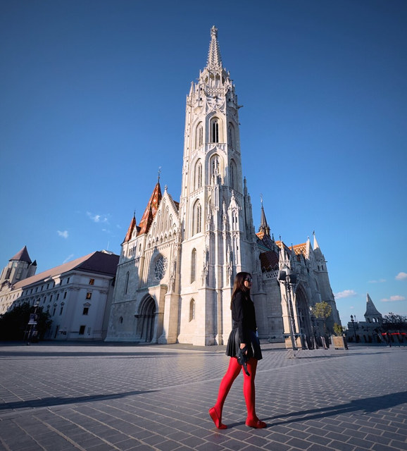 Una mujer con medias rojas cruza la plaza frente a una iglesia gótica de Budapest con tejado multicolor.