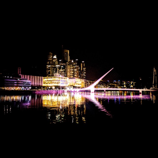 Un puente peatonal iluminado cruza el río ante los rascacielos nocturnos de Buenos Aires y sus reflejos.