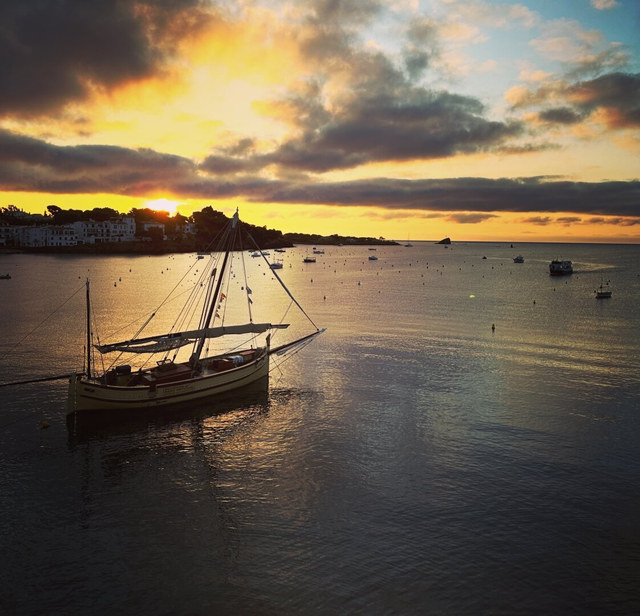Un velero tradicional fondea en la bahía de Cadaqués mientras el sol desciende entre nubes doradas.
