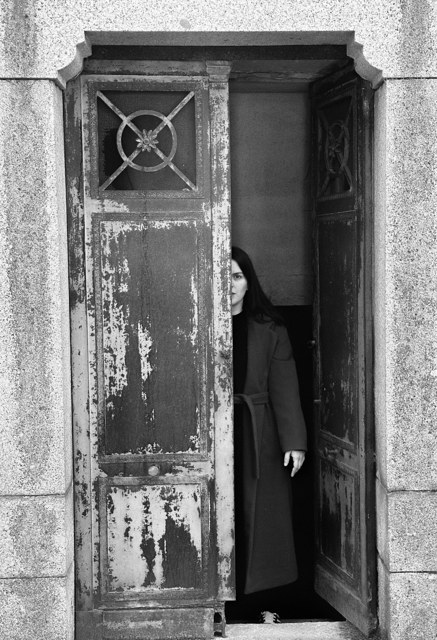 Una persona de cabello oscuro asoma detrás de la puerta metálica entreabierta y desconchada de un panteón.