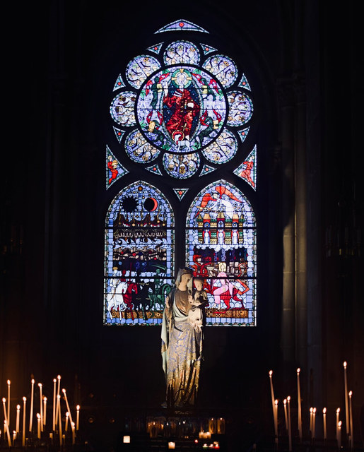 Una estatua religiosa se alza entre velas encendidas ante una vidriera policromada en una iglesia oscura.
