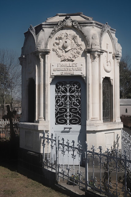 Un mausoleo blanco con relieves y puerta de hierro se levanta tras una verja ornamentada.