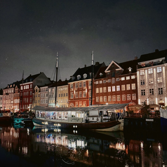 Barcos amarrados frente a casas estrechas iluminadas proyectan reflejos sobre el canal nocturno de Copenhague.