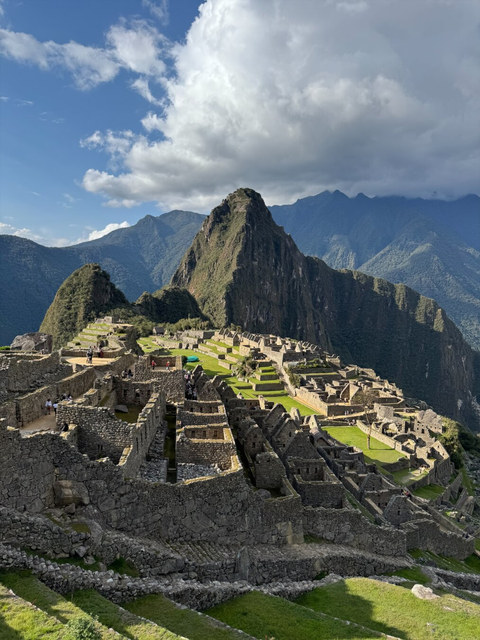 Las ruinas escalonadas de Machu Picchu se extienden bajo montañas verdes y una gran nube blanca.