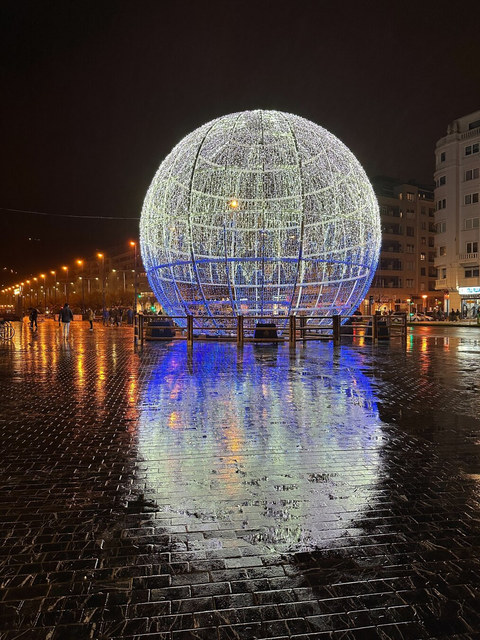 Una esfera gigante de luces blancas y azules se refleja sobre el pavimento mojado de una plaza nocturna.
