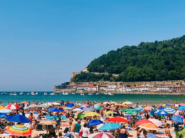 Sombrillas multicolores y bañistas llenan la playa de San Sebastián, frente a una bahía con barcos.