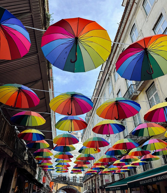 Decenas de paraguas multicolores cuelgan sobre una calle estrecha de Lisboa entre balcones y restaurantes.