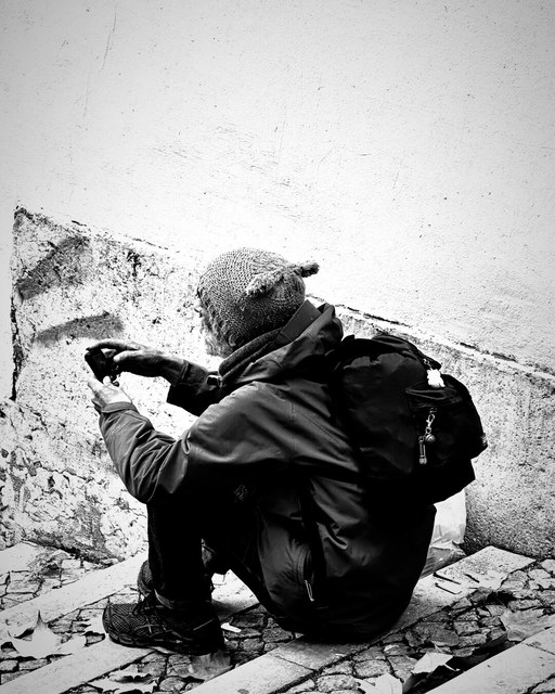 Un hombre agachado sostiene una cámara junto a una pared descascarillada, con mochila y gorro de lana.