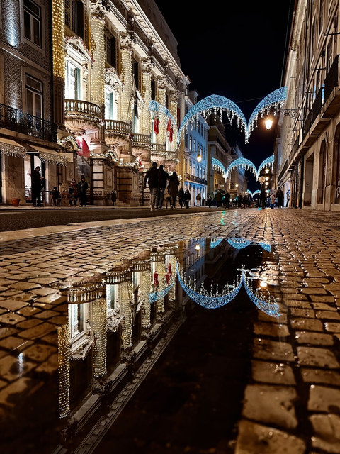 Luces festivas adornan una calle mojada de Lisboa y se reflejan en un charco entre los adoquines.