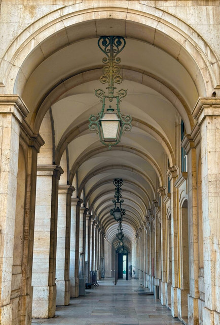 Una sucesión de arcos de piedra y faroles verdes se pierde al fondo de una galería vacía.