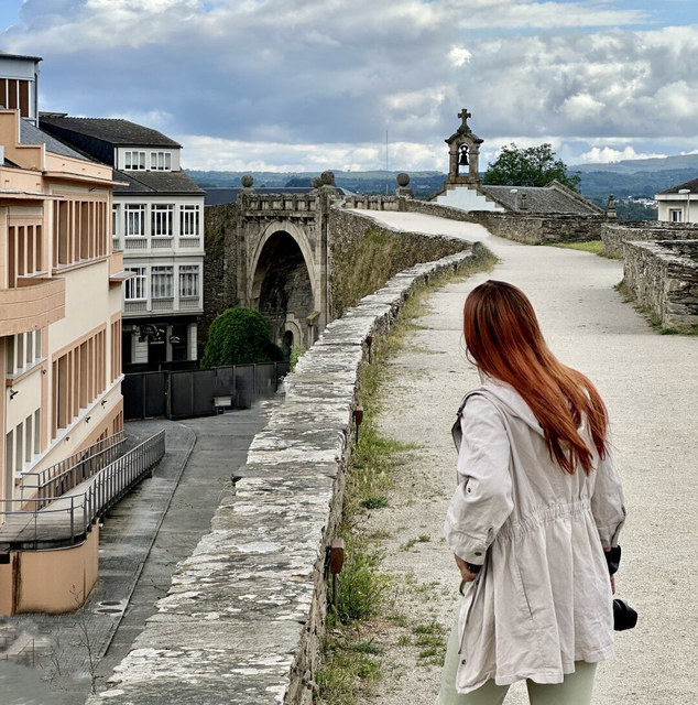 Una mujer pelirroja camina por la muralla de Lugo hacia una pequeña espadaña de piedra.