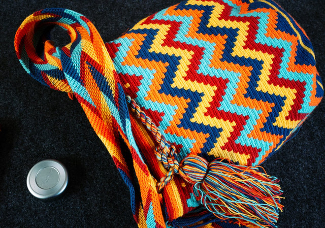 Un bolso tejido con zigzags multicolores y borla descansa sobre tela negra junto a una tapa de objetivo.