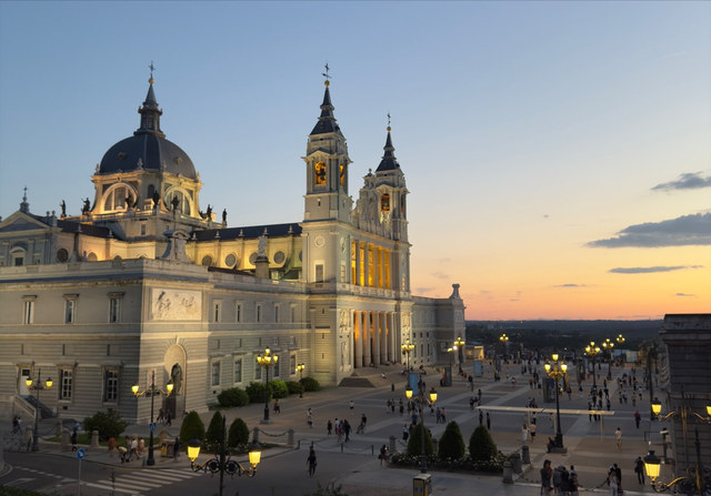 La catedral de la Almudena iluminada domina una plaza concurrida bajo un cielo rosado de Madrid.