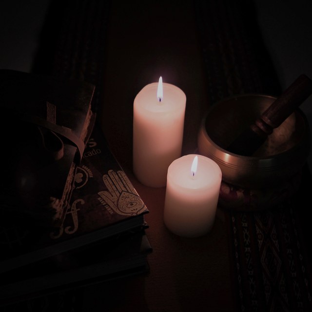 Dos velas encendidas iluminan varios libros decorados y un cuenco metálico con mazo sobre una mesa oscura.