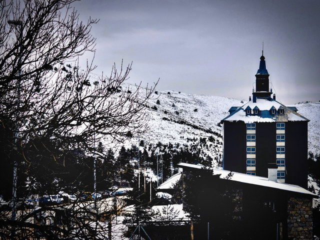 Un edificio de montaña con torre central se eleva entre tejados nevados, pinos oscuros y una ladera blanca.