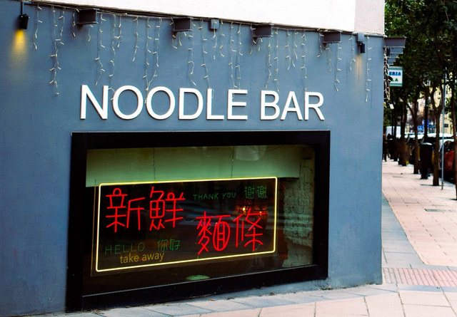 La fachada azul de un Noodle Bar incorpora un neón rojo con caracteres chinos y mensajes de comida para llevar.
