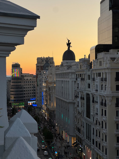 La Gran Vía de Madrid desciende entre fachadas blancas, la cúpula del Metrópolis y letreros luminosos al atardecer.