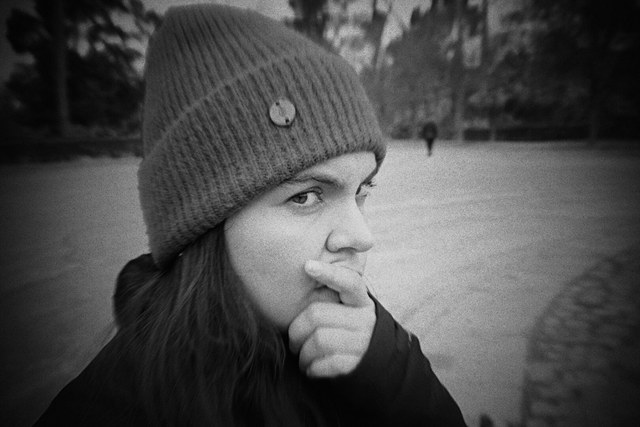 Una mujer con gorro de lana mira a la cámara mientras apoya la mano sobre la boca, en blanco y negro.