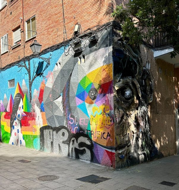 Mural geométrico multicolor y escultura de materiales reciclados cubren la esquina de un edificio de ladrillo en Madrid.
