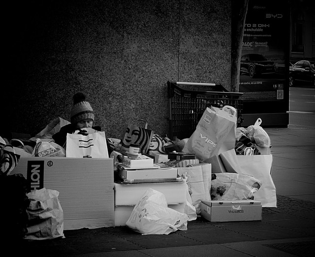 Una persona con gorro asoma entre cajas y bolsas apiladas junto a un carrito, en blanco y negro.