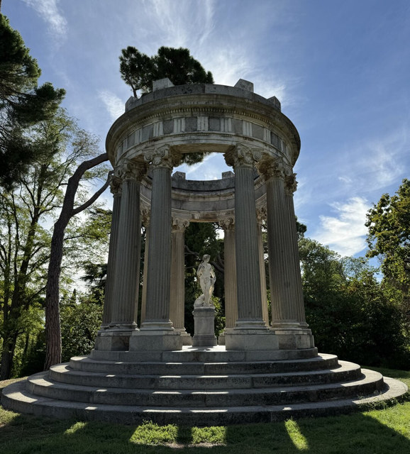 Un templete circular de columnas corintias rodea una estatua blanca entre árboles de un parque de Madrid.
