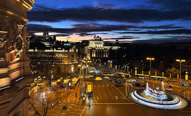 El tráfico nocturno rodea la fuente de Cibeles en Madrid, bajo edificios iluminados y largas nubes azuladas.