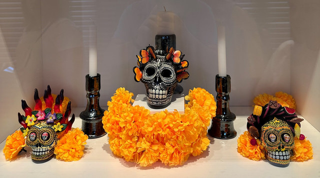 Tres calaveras decoradas con mariposas y flores descansan entre cempasúchiles naranjas y velas negras.