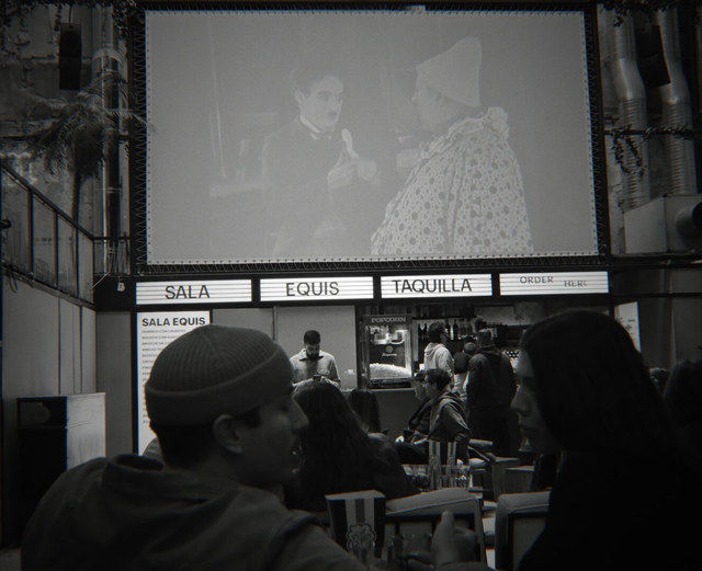 Varias personas conversan bajo una pantalla que proyecta a Chaplin, frente a la taquilla iluminada de Sala Equis en Madrid.