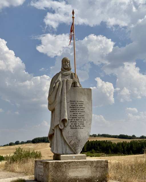 Una estatua de piedra del Cid sostiene lanza y escudo ante campos secos y grandes nubes en Mecerreyes.
