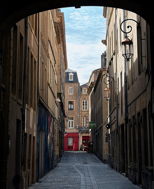 Un arco oscuro enmarca una calle estrecha de Metz con faroles y una tienda roja al fondo.