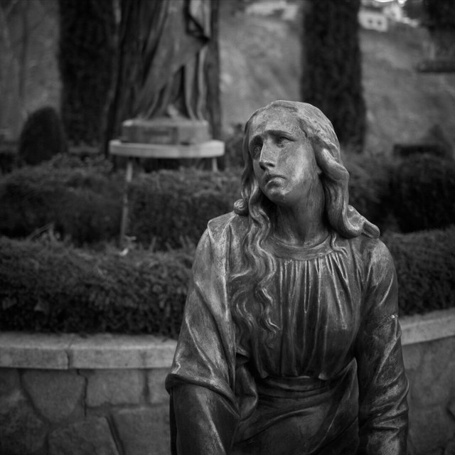 Una estatua de mujer sentada, con la mirada elevada, ocupa un jardín entre setos y monumentos funerarios.