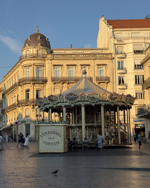 Un carrusel iluminado ocupa una plaza de Montpellier, rodeado de edificios clásicos y una paloma solitaria.