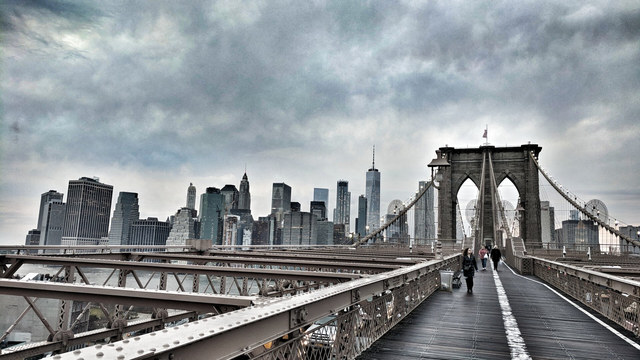 Peatones cruzan la pasarela del puente de Brooklyn hacia Manhattan bajo un cielo cubierto de nubes grises.
