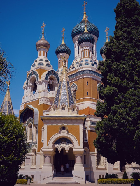 Una catedral ortodoxa de ladrillo y cúpulas bulbosas se alza entre cipreses bajo el cielo azul de Niza.