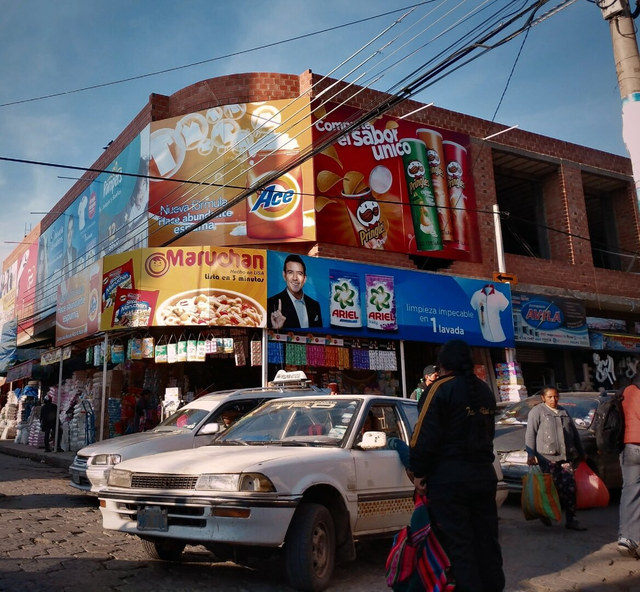 Un coche blanco y varios peatones pasan ante una fachada cubierta de anuncios comerciales en La Paz.
