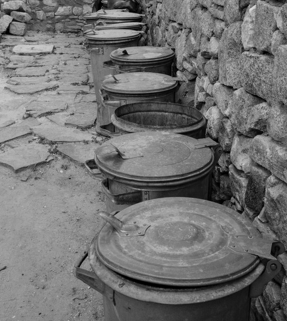 Una hilera de cubos metálicos bordea un camino empedrado, con uno abierto junto al muro de piedra.