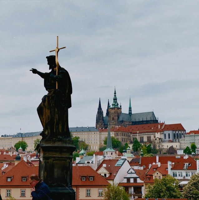 Una estatua sostiene una cruz dorada y señala sobre tejados rojos hacia la catedral de Praga.