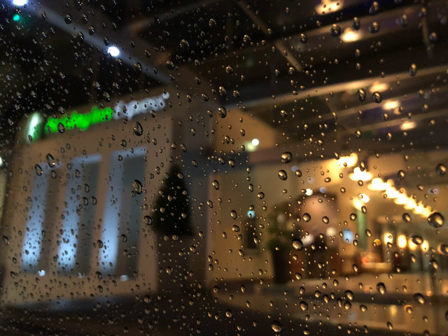 Gotas de lluvia cubren un cristal y enfocan parcialmente las luces cálidas de un interior desenfocado.