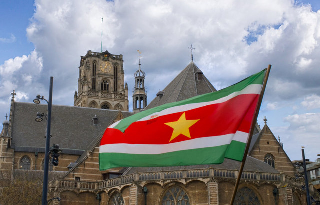 Una bandera de Surinam ondea ante una iglesia de piedra en Rotterdam bajo grandes nubes blancas.