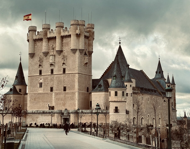 El Alcázar de Segovia combina una torre almenada y tejados puntiagudos bajo un cielo cubierto de nubes.