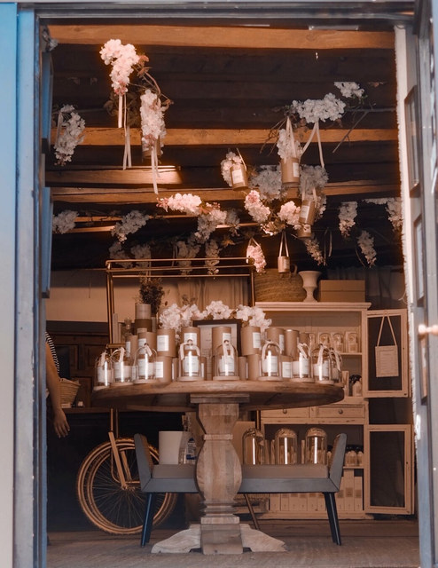 Una mesa cubierta de botellas ocupa una tienda decorada con flores colgantes y una bicicleta al fondo.