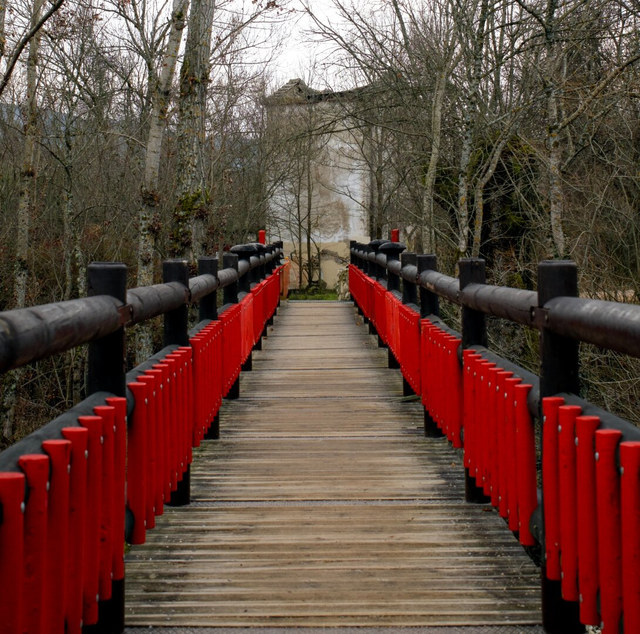 Una pasarela de madera con barandillas rojas cruza un bosque sin hojas hacia un edificio parcialmente oculto.