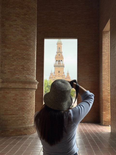Una mujer con sombrero fotografía una torre de ladrillo enmarcada por columnas y un gran vano rectangular.