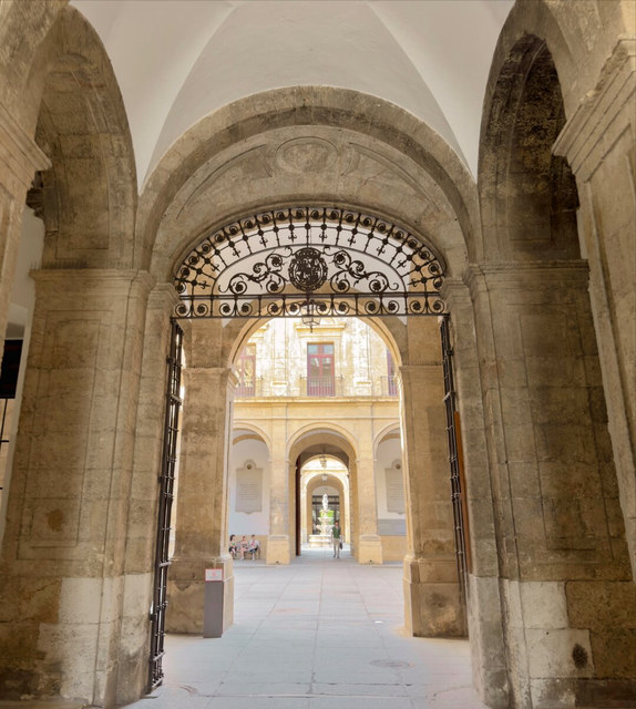 Una reja de hierro abierta enmarca un patio sevillano con arcos, balcones y visitantes al fondo.