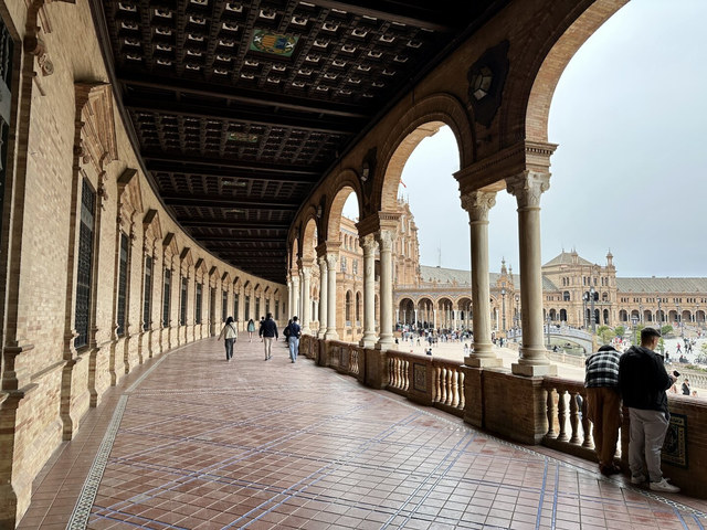 Visitantes recorren una galería curva de arcos y columnas con vistas a la Plaza de España de Sevilla.