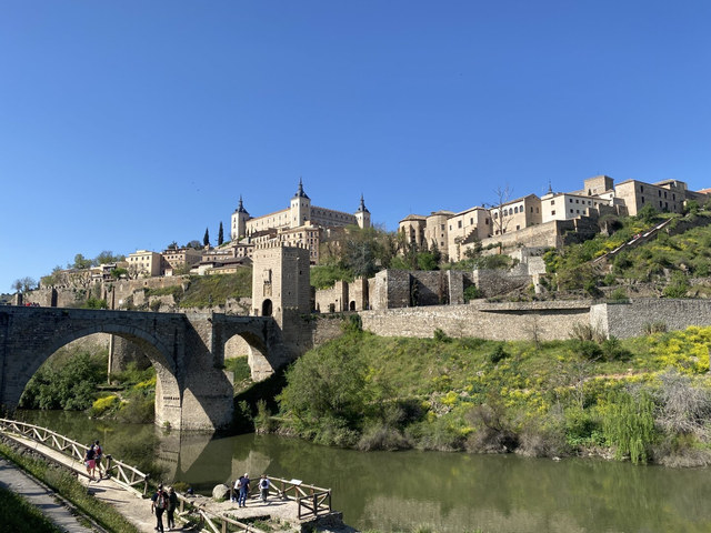 Un puente medieval cruza el río hacia las murallas y edificios históricos de Toledo bajo un cielo azul.